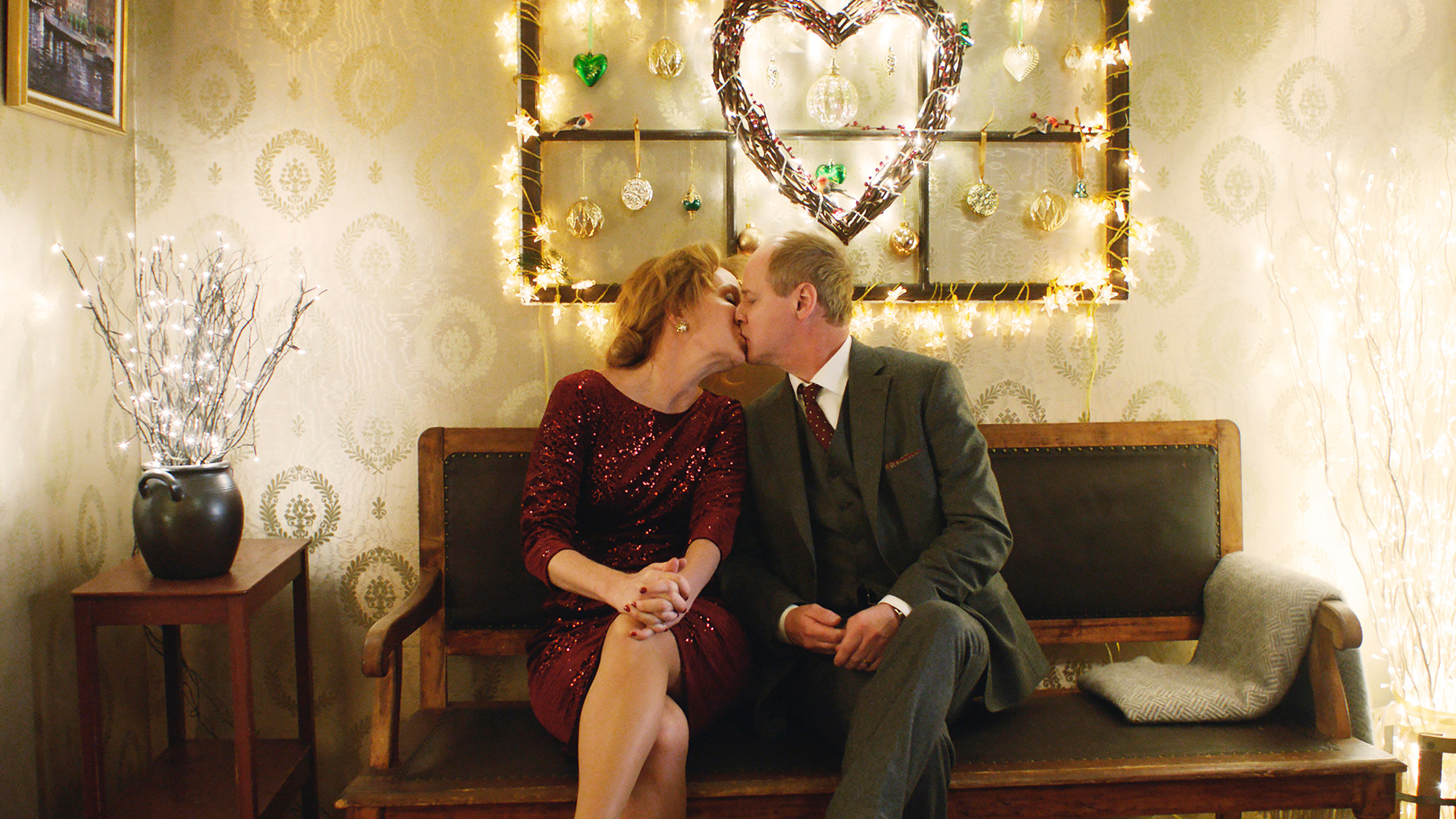 Filmstill aus "Eine schöne Bescherung": Ein sich küssendes Paar um die 60 sitzt auf einer lederbezogenen Holzbank unter einem weihnachtlich dekorierten Herz.