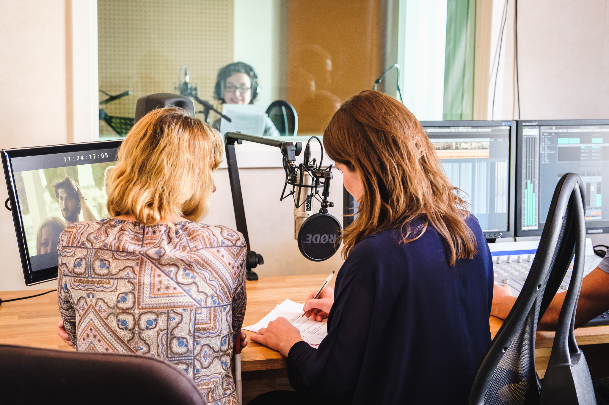 In einem Tonstudio: Im Vordergrund zwei Frauen mit dem Rücken zur Kamera. Eine der beiden notiert etwas. Im Hintergrund durch eine Scheibe eine Sprecherin bei der Aufnahme.