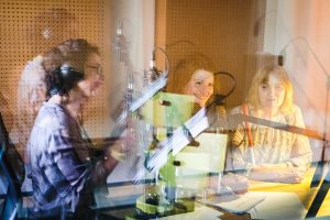 Blick durch die Glasscheibe in ein Tonstudio, in dem zwei Frauen sitzen. Eine der beiden Frauen trägt ein Langstock bei sich. In der Scheibe spiegelt sich eine Sprecherin am Mikrofon.