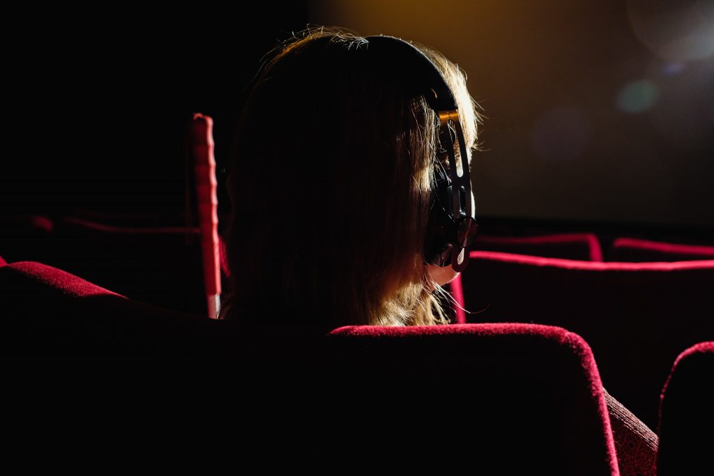 Eine Frau von hinten in einem dunklen Kinosaal. Neben ihr lehnt ein Langstock, sie trägt große Kopfhörer.