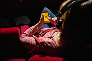 Blick über die Schulter einer Frau in einem Kinosessel. Sie trägt große Kopfhörer und hält ein Handy in der Hand, auf dem das Logo der App "Greta" zu erkennen ist.