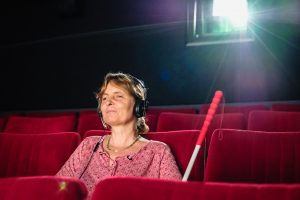 Barbara Fickert mit schwarzem Kopfhörer auf dem Kopf im Kino, ihr Langstock lehnt zu ihrer Linken