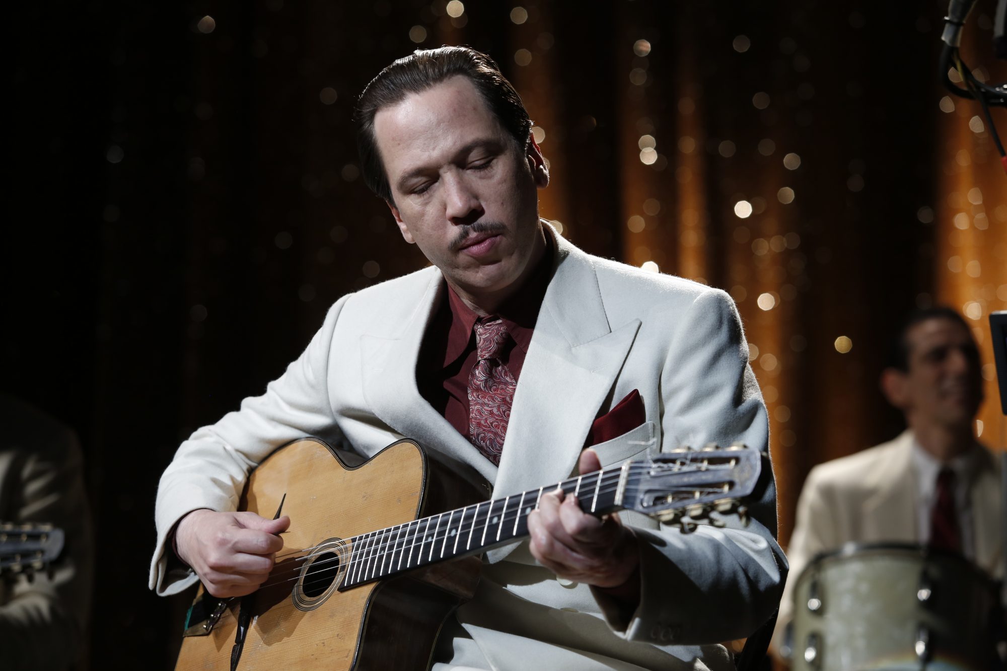 Filmstill aus "Django - Ein Leben für die Musik": Django Reinhardt mit dunklen, zurückgegelten Haaren und und schmalem Schnurrbart an der Gitarre. Er trägt ein weißes Sakko über einem weinroten Hemd mit farblich passendem Einstecktuch in der Brusttasche. Hinter ihm ein Schlagzeuger vor einem gold-glitzernden Vorhang.