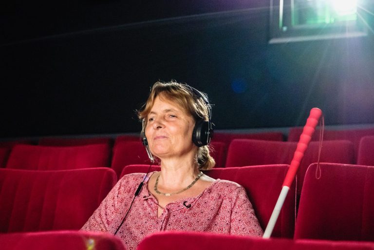Barbara Fickert mit schwarzem Kopfhörer auf dem Kopf im Kino, ihr Langstock lehnt zu ihrer Rechten.