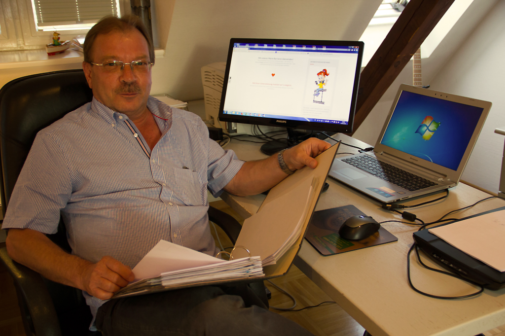 Ein Mann mittleren Alters am Schreibtisch mit Monitor und Laptop, er hält einen Aktenordner in den Händen