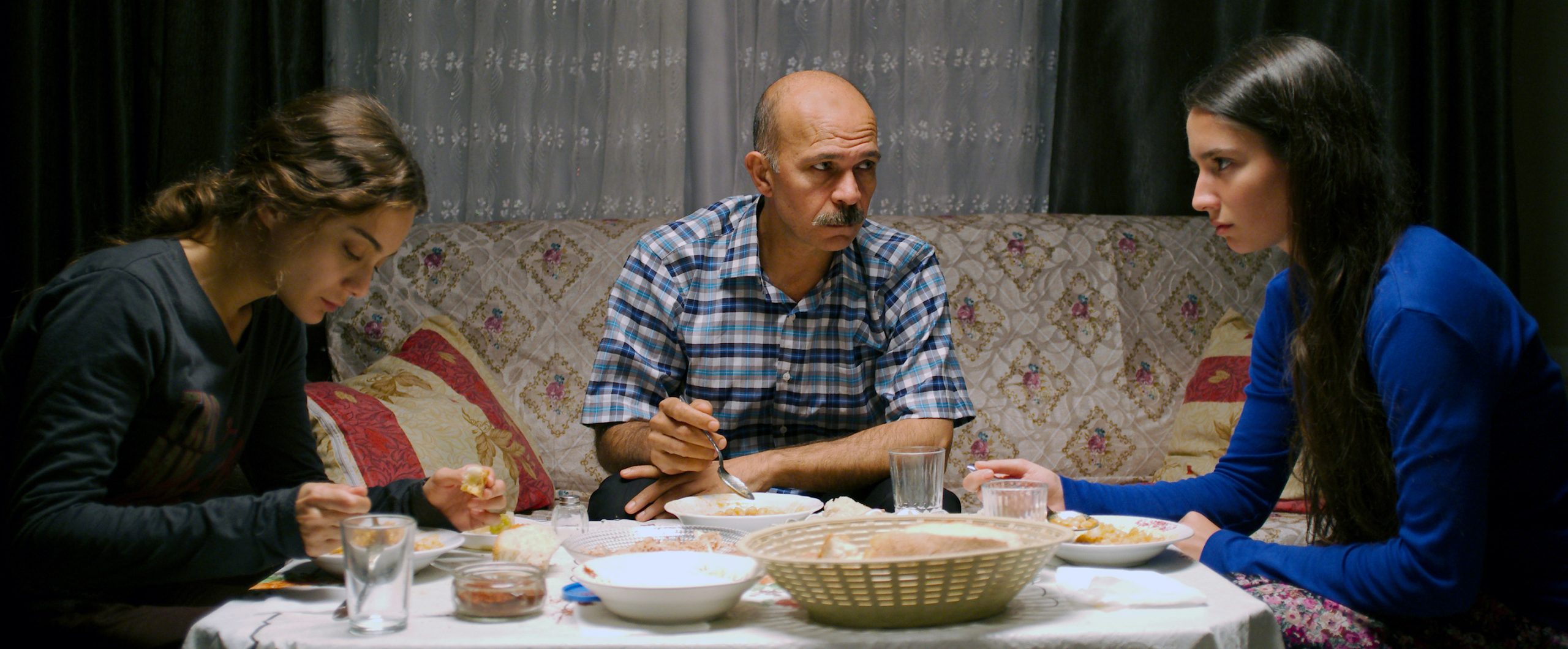 Filmstill aus dem Film "Sibel": Zwei junge Frauen und ein älterer Mann sitzen in einem Wohnzimmer beim Essen.