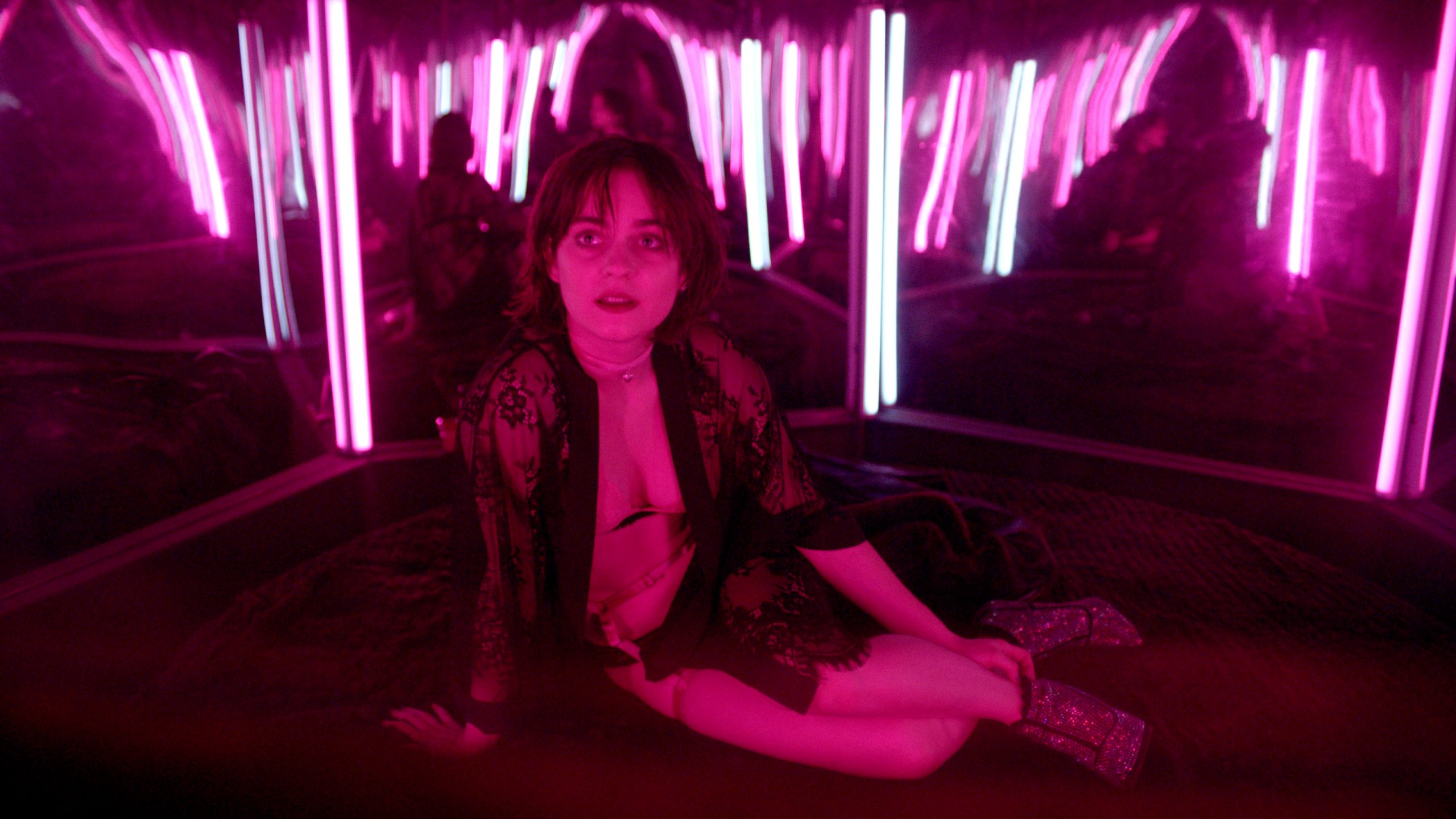 Filmstill aus "O Beautiful Night": Eine leicht bekleidete Frau liegt lassiv auf einem runden Bett in einem rot beleuchteten Spiegelzimmer.