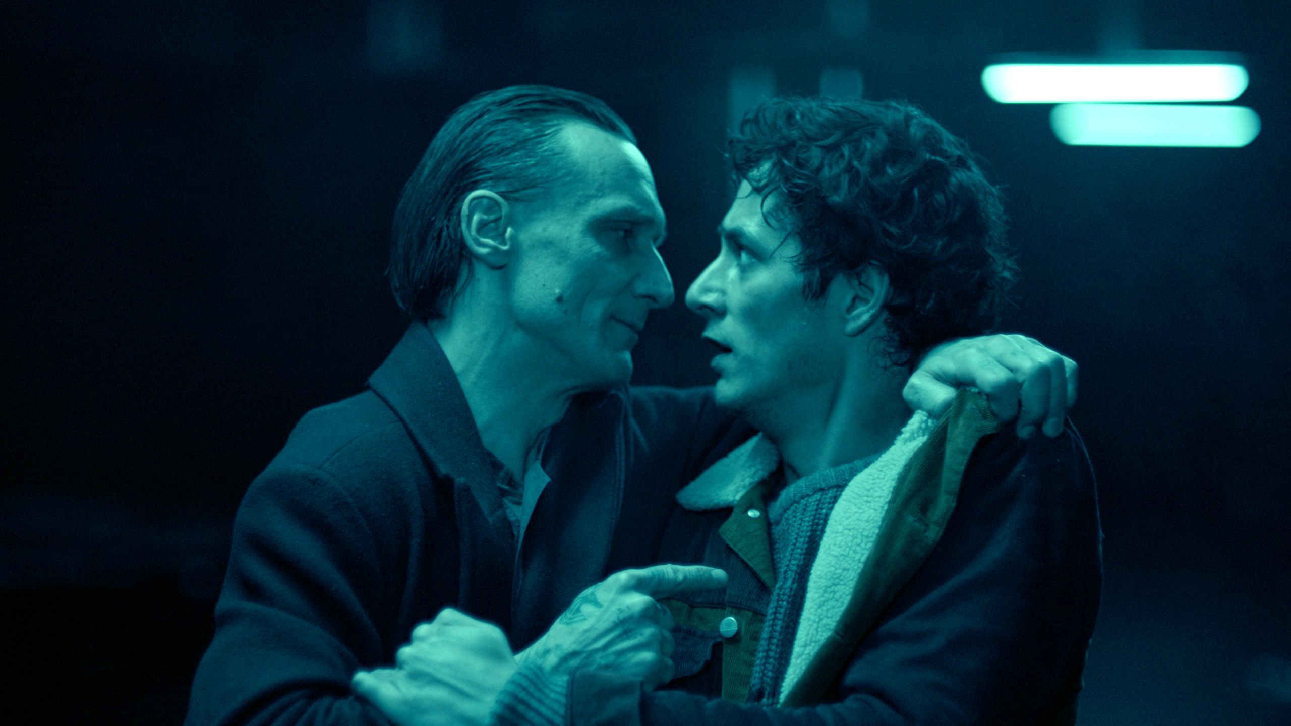 Filmstill aus "O Beautiful Night": In blau gedämmten Licht zwei Männer. Einer hat den Arm um den anderen gelegt, der andere schaut erschrocken.