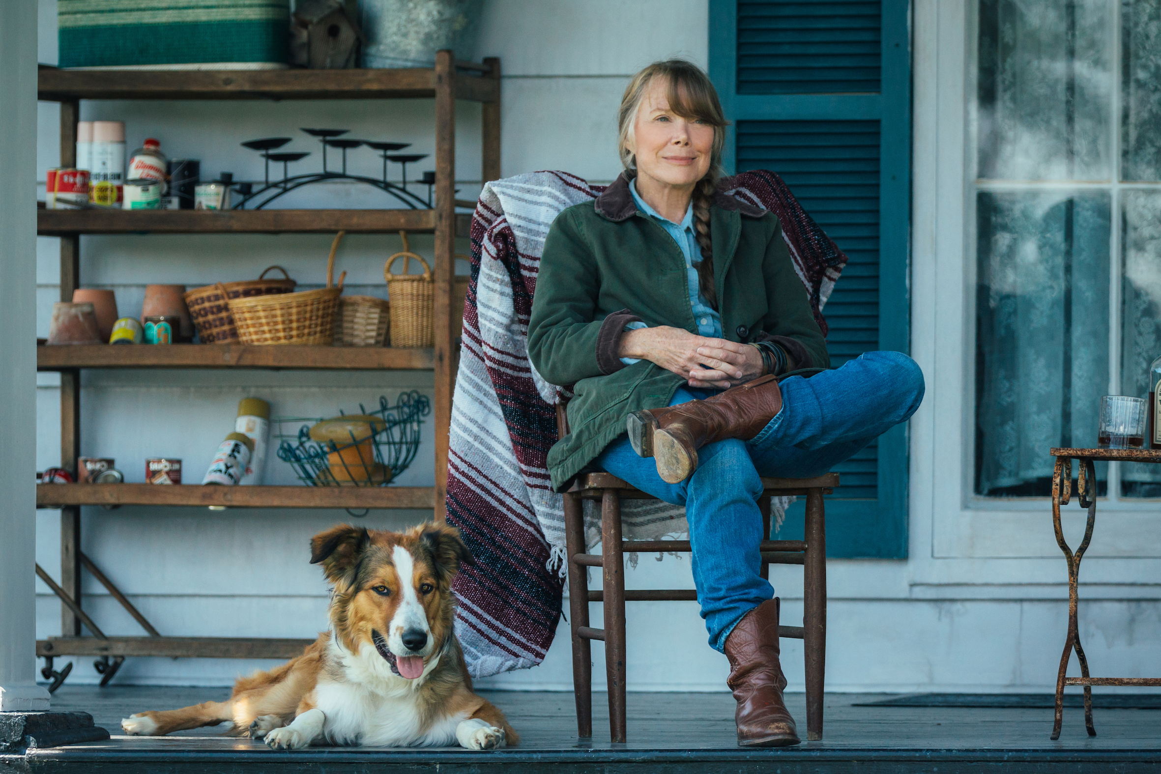 Filmstill aus "Ein Gauner & Gentleman": Eine Frau mittleren Alters sitzt lässig auf der Terrasse vor einem Haus. Sie trägt Westernstiefel, neben ihr ein Hund mit weißer Schnauze.