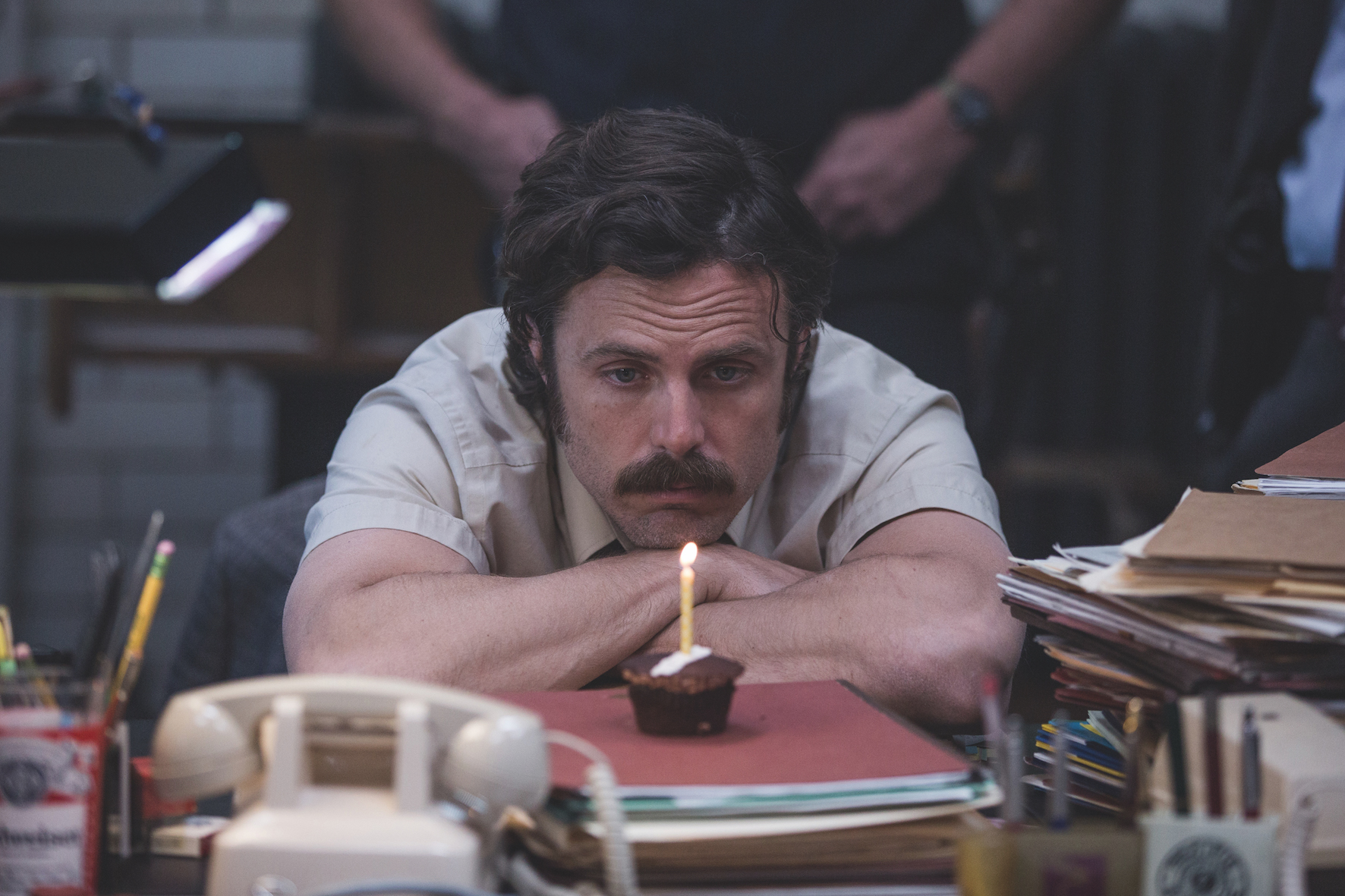 Filmstill aus "Ein Gauner & Gentleman": Ein Mann mittleren Alters sitzt in einem Büro und schaut betrübt auf einen Brownie mit brennender Kerze.