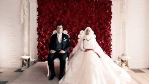 Filmstill von "Nur eine Frau": Ein starr nebeneinander sitzendes Brautpaar vor einer Rosenwand.