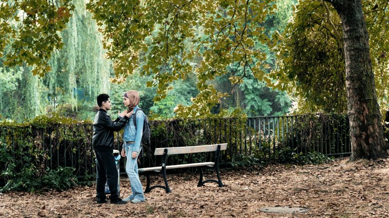 Filmstill von "Nur eine Frau": Eine junge Frau mit Kopftuch und ein junger dunkelhaariger Mann mit Lederjacke an einer Parkbank am See.