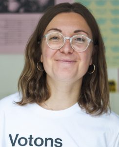 Eine strahlende junge Frau mit heller Hornbrille und schulterlangem braunem Haar