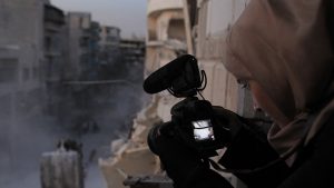 Eine junge Frau mit Kopftuch filmt aus einem Fenster auf eine Straße mit zerstörten Gebäuden.