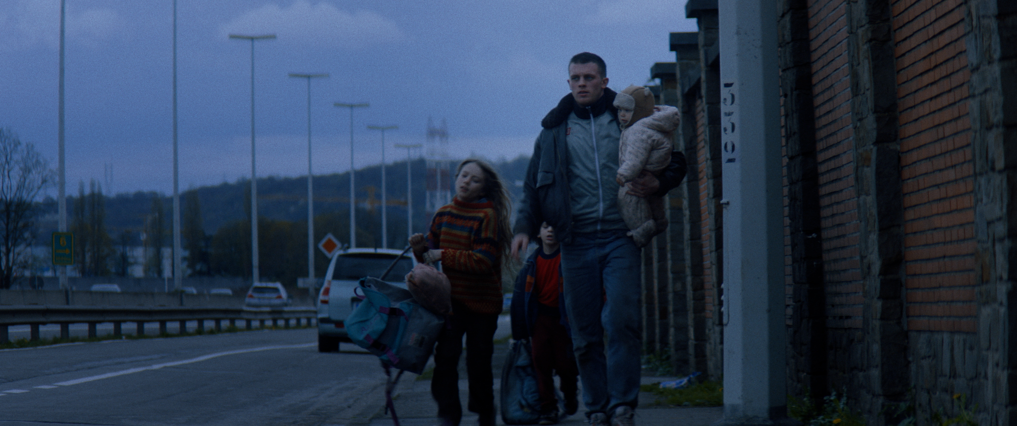 Filmstill aus "Kids Run": Ein Mann mit drei Kindern eilt in der Dämmerung am Rand einer Schnellstraße entlang.