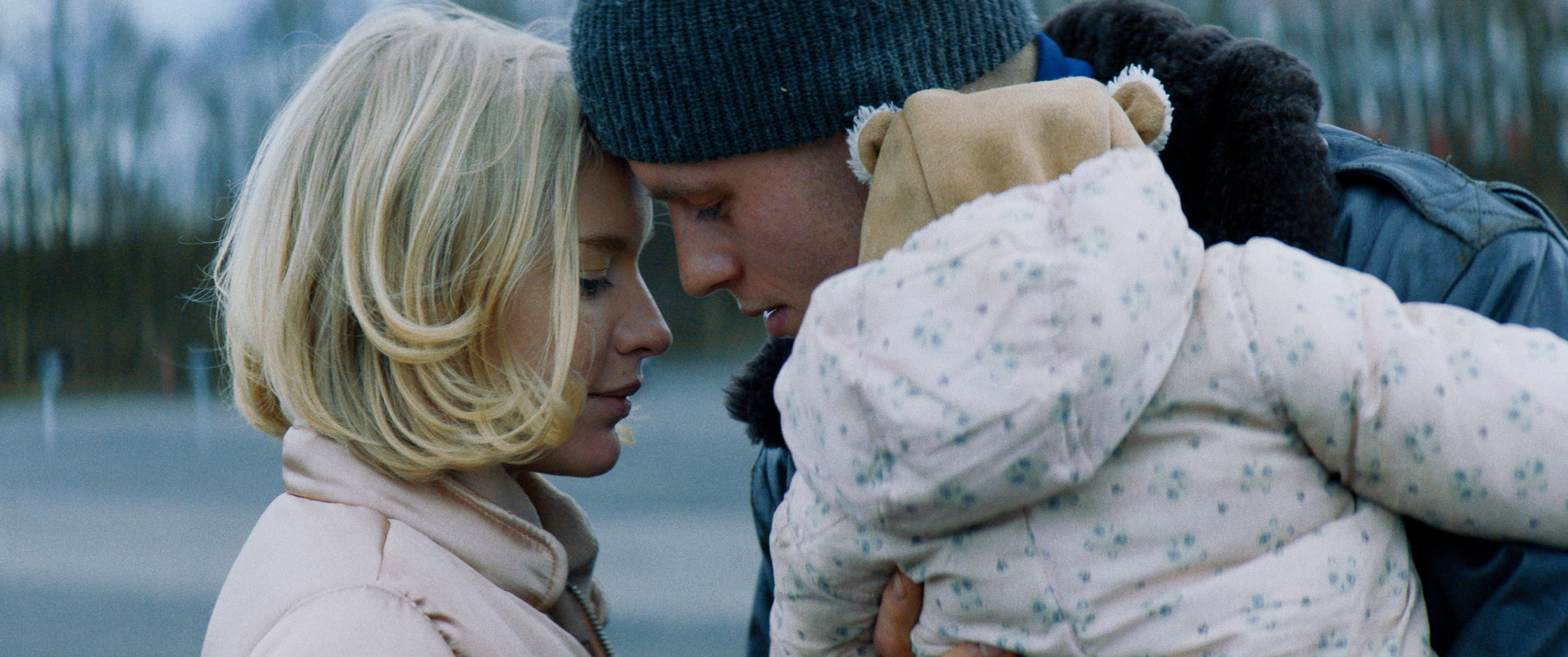 Filmstill aus "Kids Run": Ein Mann und eine Frau einander Kopf an Kopf zugewandt, der Mann trägt ein Baby mit Winterjacke und Fellmütze auf dem Arm.
