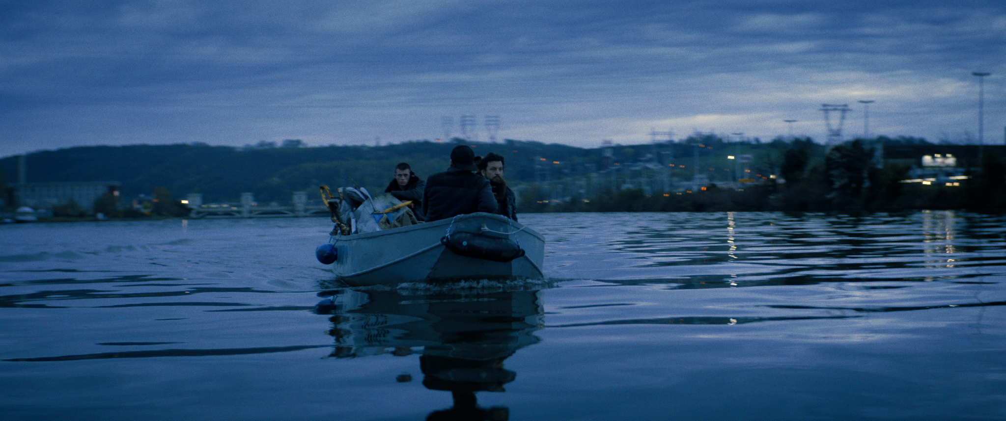 Filmstill aus "Kids Run": Im Dämmerlicht mehrere Männer in einem Boot auf dem Wasser, im Hintergrund beleuchtete Häuser und Strommasten in der hügeligen Landschaft.