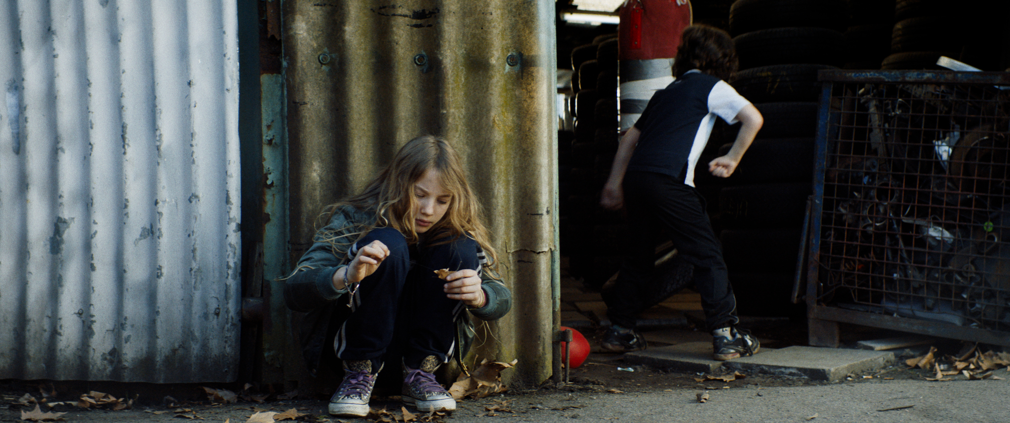 Filmstill aus "Kids Run": Ein Kind mit langem, wallendem Haar hockt am Boden vor einer Wellblechwand. An seiner Hand deutet sich eine baumelnde Handschelle an. Im Hintergrund spielt ein anderes Kind.