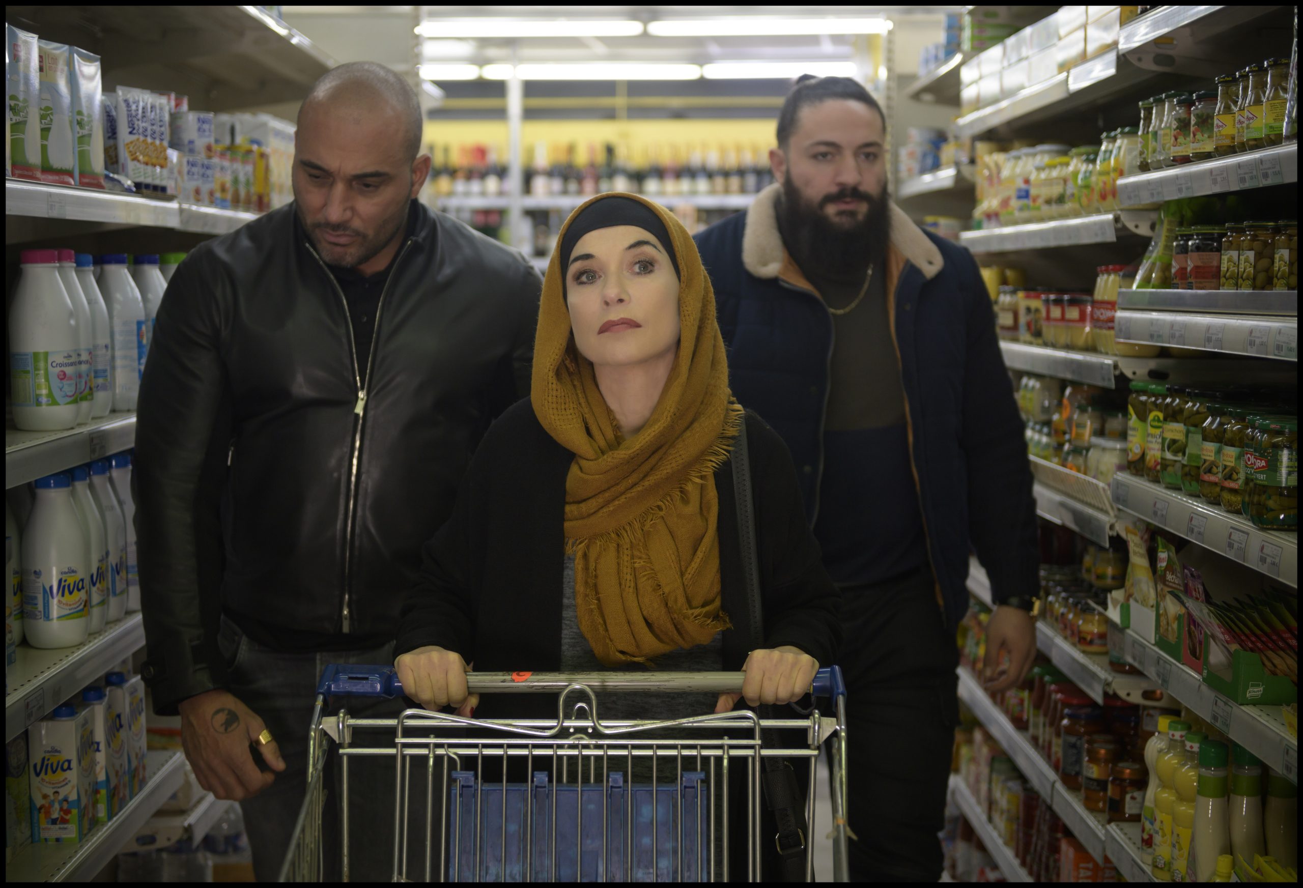 Eine dunkel geschminkte Frau mit Kopftuch, begleitet von zwei südländisch aussehenden Männern, schiebt einen Einkaufswagen durch einen Supermarkt.