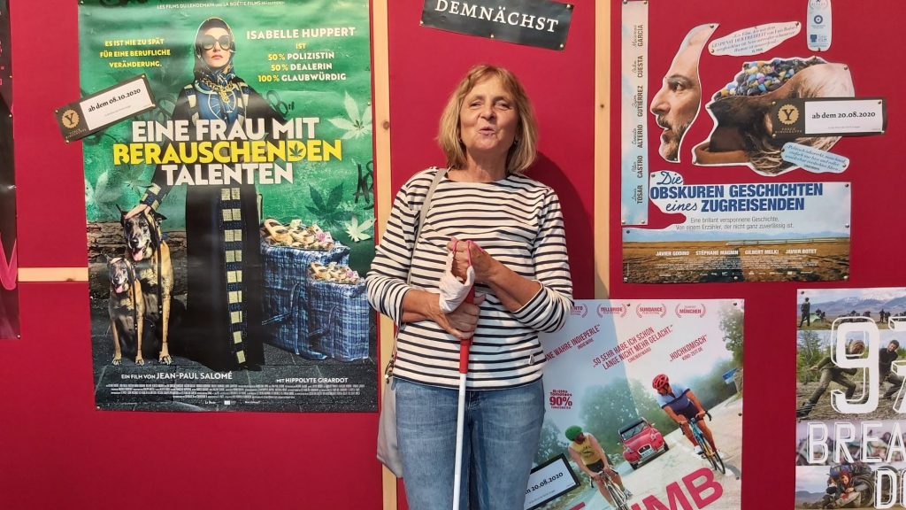 Die Blindgängerin steht im Foyer eines Kinos. Sie trägt Jeans und ein gestreiftes Shirt, in den Händen Langstock und Maske. Sie schaut fröhlich in die Kamera. Neben ihr an der Wand das Filmplakat zu "Eine Frau mit berauschenden Talenten".