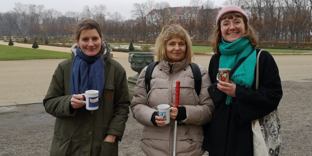Lena, Barbara und Anna stehen nebeneinander im Schloßpark Charlottenburg. Sie halten Tassen in den Händen und lachen in die Kamera. Außer den dreien ist kein Mensch zu sehen.