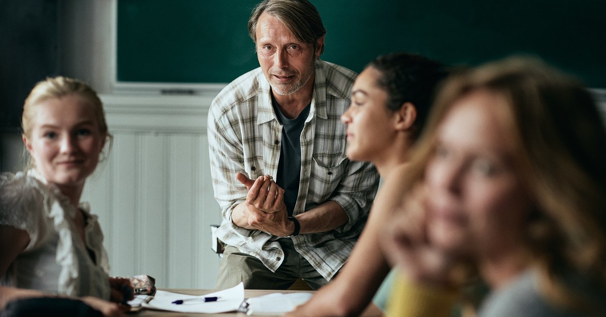 Der Lehrer Martin vor einer Schulklasse. Er hat die Hände ineinandergelegt und scheint freundlich, aber eindringlich zu einem Schüler zu sprechen. Zwei Mädchen aus der vorderen Reihe schauen hinter sich.