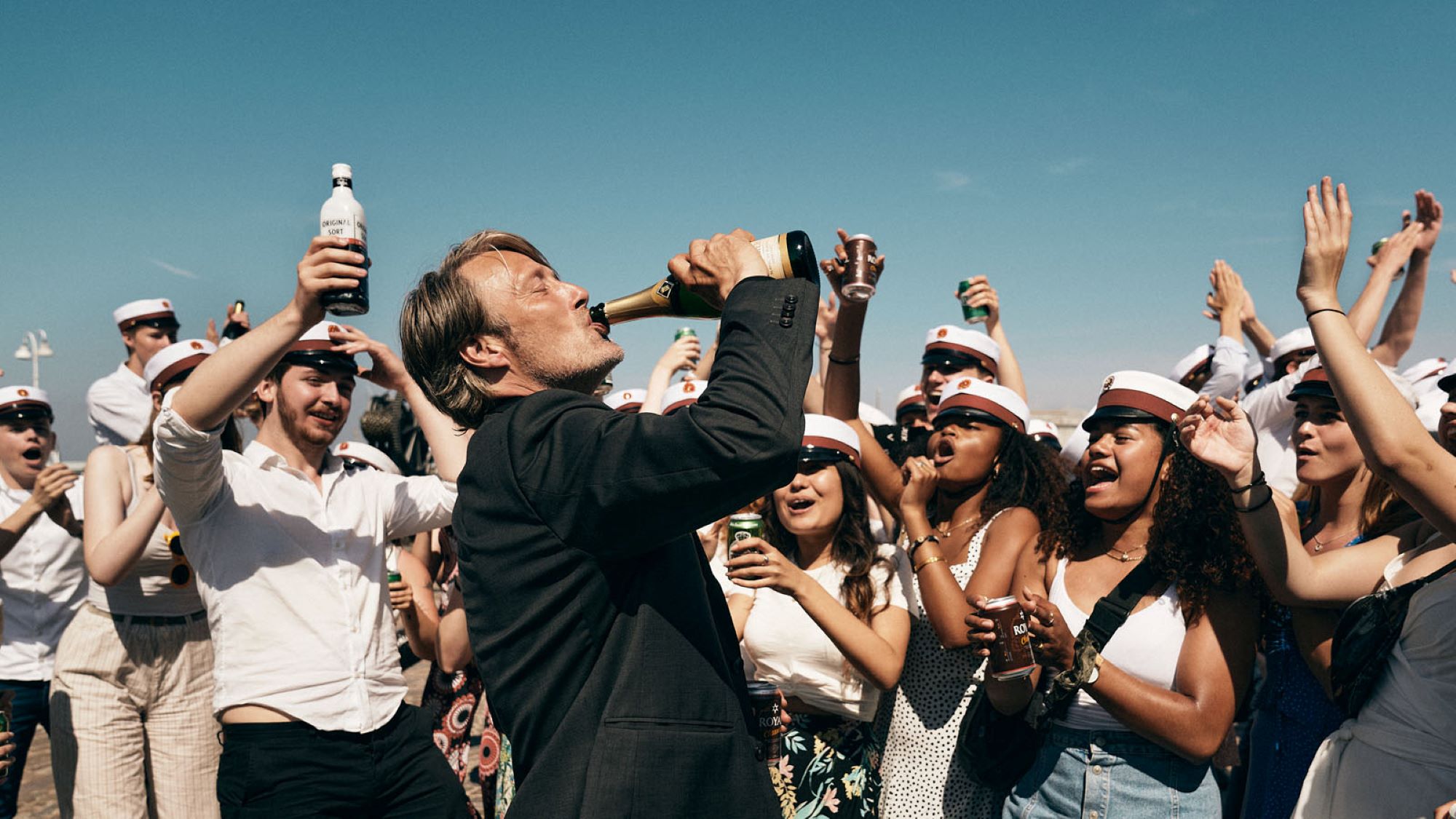 Mads Mikkelsen, der Hauptdarsteller des Films "Der Rausch", trinkt aus einer Sektflasche. Er ist umringt von einer Gruppe jubelnder Abiturienten, die weiße Schulmützen tragen. Viele halten Flaschen oder Dosen in der Hand und prosten ihrem Lehrer zu.