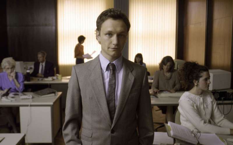 Der Hauptdarsteller Daniel Michel als junger Banker Rüdi in einem Großraumbüro. Er trägt Hemd mit Schlips und Sakko sowie ein verschmitztes Lächeln.