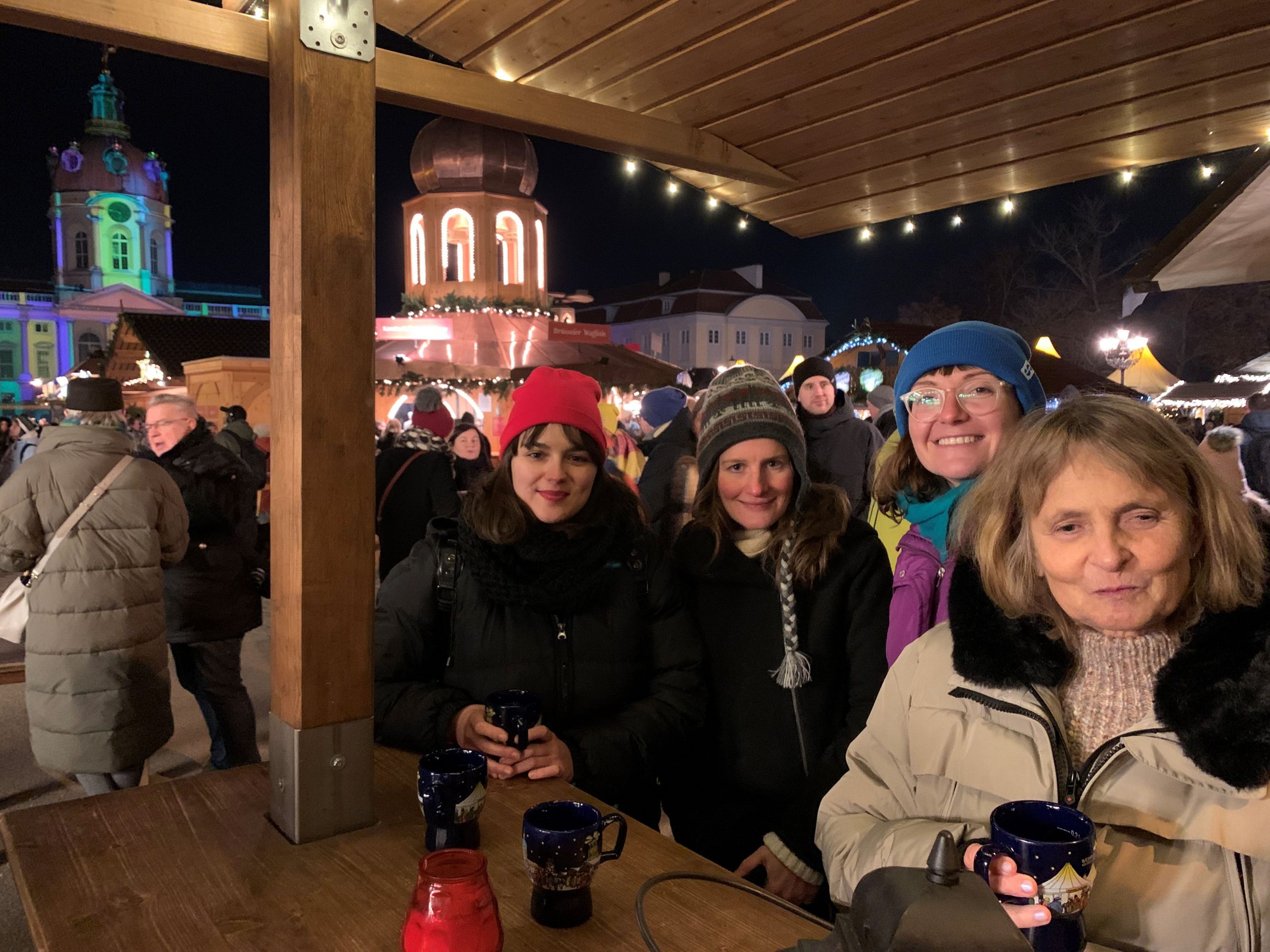 Auf dem Weihnachtsmarkt am Schloss Charlottenburg, im Hintergrund der beleuchtete Turm des Schlosses. In dicken Winterjacken stehen Danae, Lena, Anna und Barbara mit Glühweinbechern an einem Holztisch.