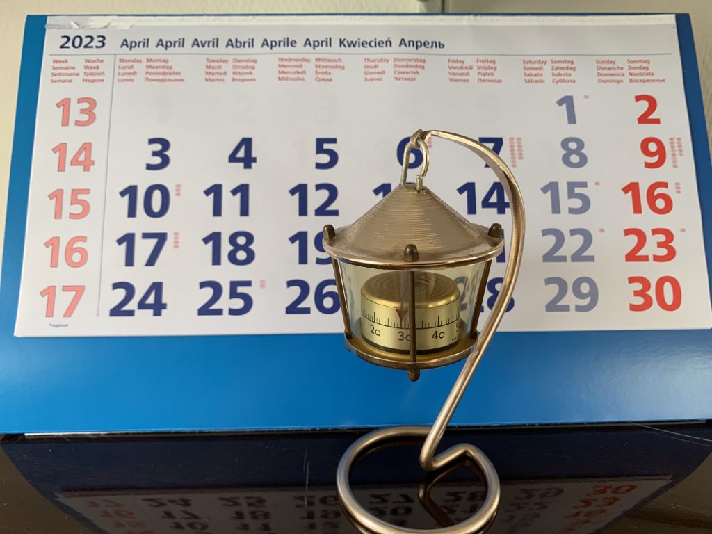 Vor einem Kalenderblatt des Monats April 2023 steht ein altes Zimmerthermometer aus Messing in Form einer Laterne. Der Zeiger steht auf 29 Grad.