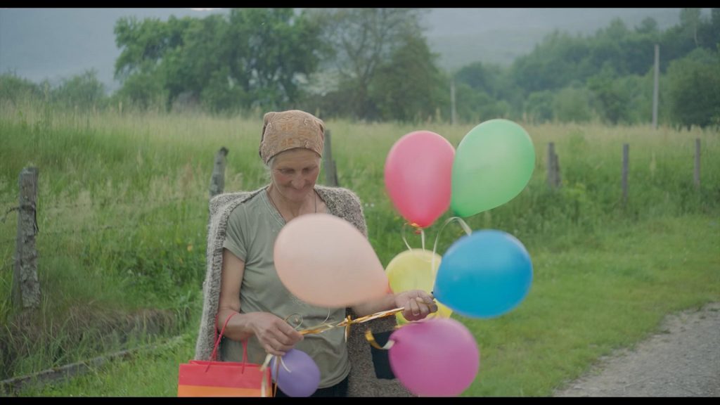 Eine ältere Frau mit Kopftuch steht vor einer hohen Wiese, die mit einfachen Holzpfählen und Gitterdraht eingezäunt ist. Die Frau hält sieben bunte Luftballons an einem Band, an ihrem rechten Arm hängt eine bunte Geschenktüte. An der linken Hand trägt sie einen Ring, ihr Blick ist gesenkt.