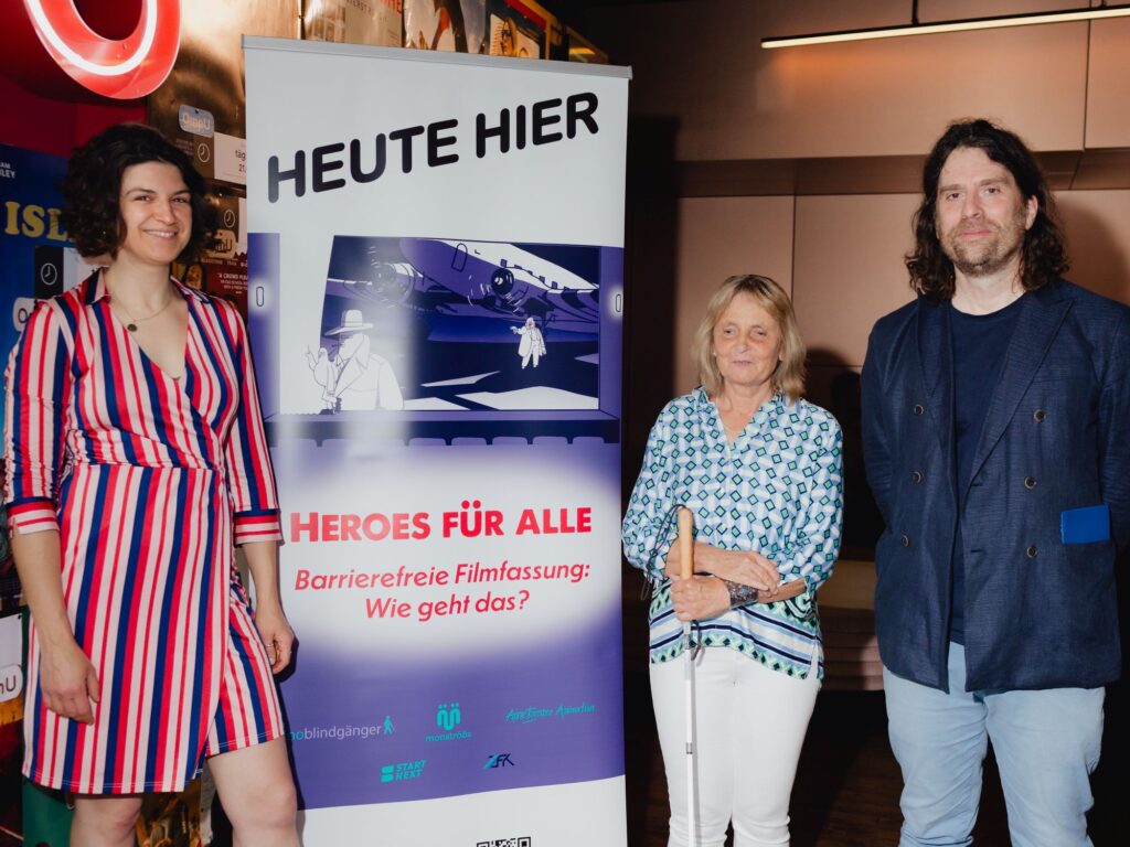 Im Foyer des Kinos Delphi LUX. In der Bildmitte das Banner mit dem Filmplakat "Heroes für alle". Links daneben steht Anne Isensee, rechts daneben stehen Barbara Fickert und Ralf Krämer.