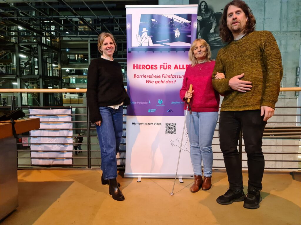 Vor dem Geländer einer Galerie stehen Stefanie Georgi, die Blindgängerin und Ralf Krämer. Zwischen ihnen ein Aufsteller mit dem Filmplakat "Heroes für alle". Dahinter das hohe Atrium im modernen Gebäude der Filmuniversität Babelsberg Konrad Wolf mit Sichtbeton und offenen Stahlträgern.