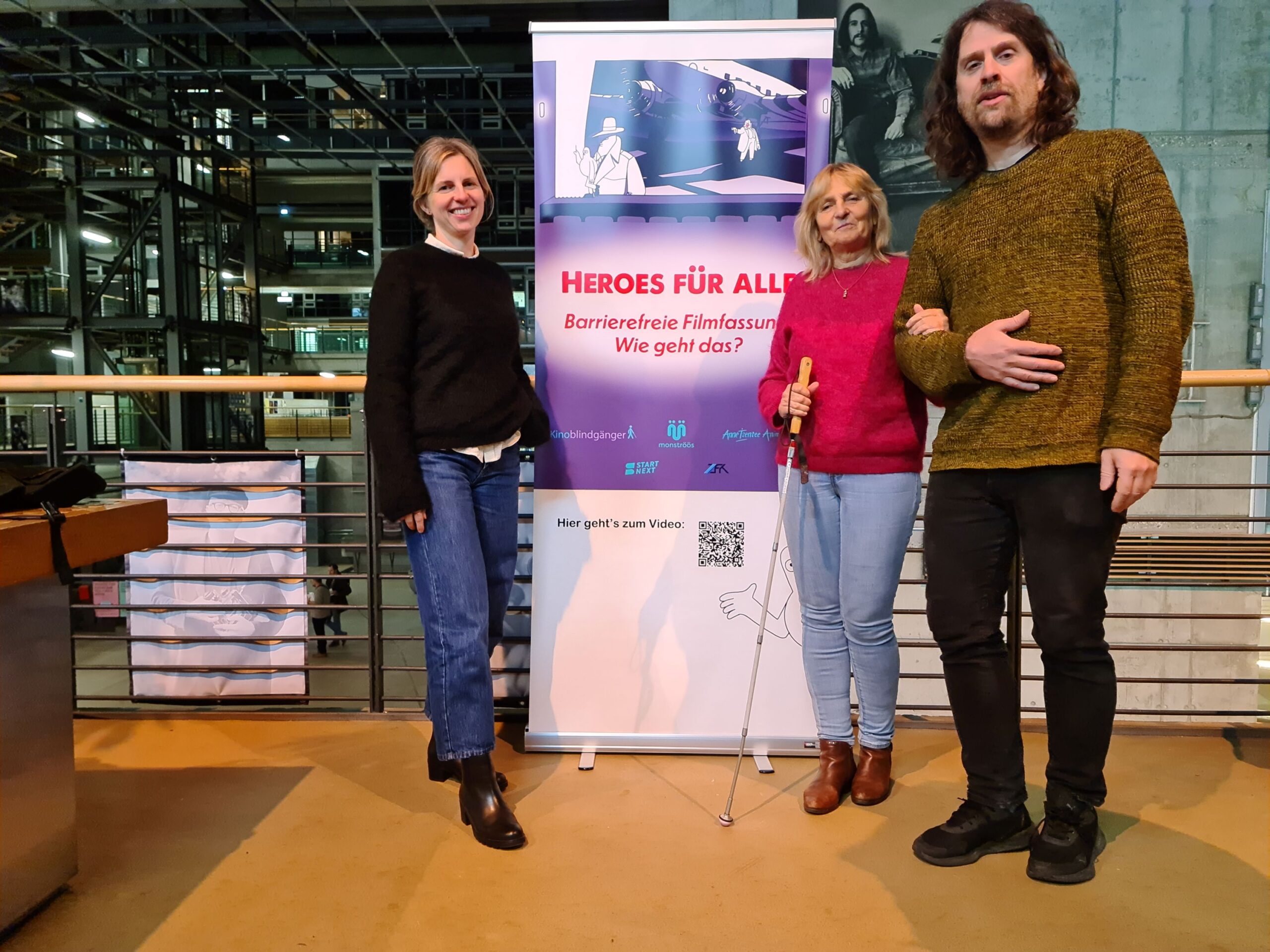 Vor dem Geländer einer Galerie stehen Stefanie Georgi, die Blindgängerin und Ralf Krämer. Zwischen ihnen ein Aufsteller mit dem Filmplakat "Heroes für alle". Dahinter das hohe Atrium im modernen Gebäude der Filmuniversität Babelsberg Konrad Wolf mit Sichtbeton und offenen Stahlträgern.