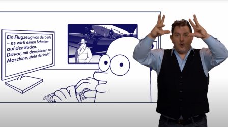 Auf der linken Bildhälfte in einem blauen feinen Rahmen eine Filmszene aus „HEROES FÜR ALLE“. Luca sitzt am Computer und schaut uns über die Schulter an. Auf dem Monitor vor ihm entsteht der Text einer Audiodeskription: "Ein Flugzeug von der Seite..." Auf dem Monitor daneben, halb von Lucas Kopf verdeckt: Die Szene aus "Heroes für alle..." mit dem Helden auf dem Rollfeld. Am rechten Bildrand steht der Gebärden-Dolmetscher Jan Sell. Er ist Ende 30, hat verstrubbeltes, dunkles Haar und einen Dreitage-Bart. Er trägt eine dunkle Weste über einem hellblauen Hemd. Mit großen Augen schaut er uns an, hält die Hände auf Kopfhöhe und spitzt die Lippen.